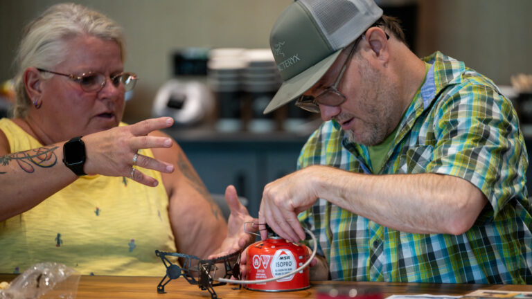 A man and a woman are working together at a table, with the man focused on a red gas canister while the woman gestures with her hands.