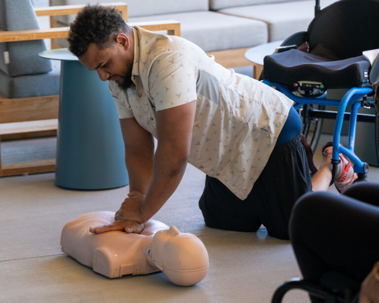 A man performs CPR on a training mannequin while kneeling on the floor in a modern indoor setting.