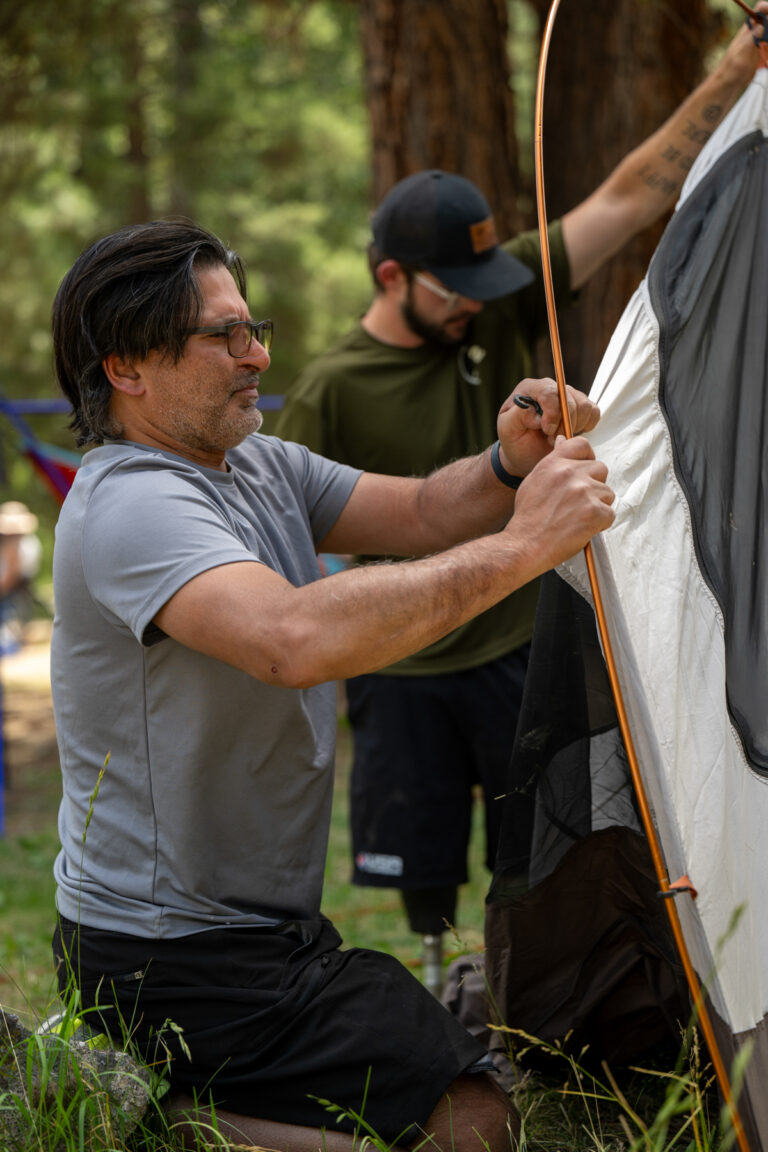 Two individuals are setting up a tent in a wooded area, with one man kneeling and adjusting a tent pole while the other assists from the background.