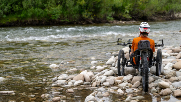A person in an all-terrain wheelchair sits on a rocky riverbank, facing a flowing river, wearing a helmet and an orange shirt.