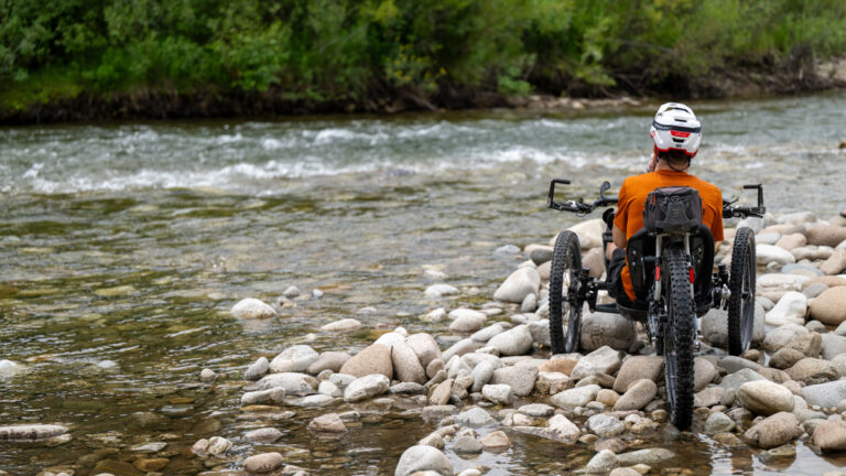 A person in an all-terrain wheelchair sits on a rocky riverbank, facing a flowing river, wearing a helmet and an orange shirt.