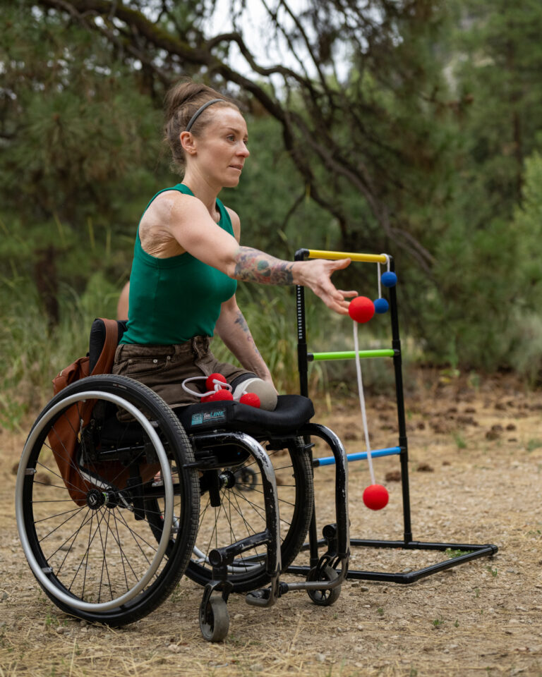 A woman in a green tank top sits in a black wheelchair, preparing to throw a red ball towards a colorful game structure in an outdoor setting filled with trees.