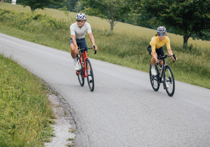 Two cyclists riding road bikes on a paved rural road bordered by grass fields and trees during a training ride.