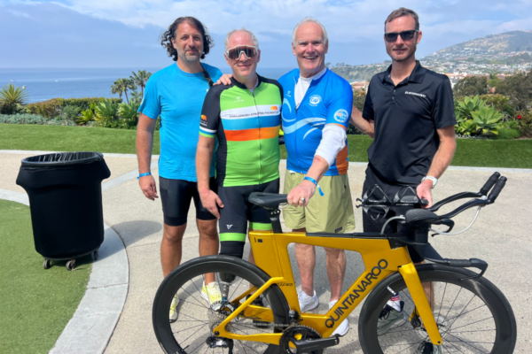 Four people standing outdoors beside a yellow Quintana Roo bicycle with an ocean and coastal landscape in the background.