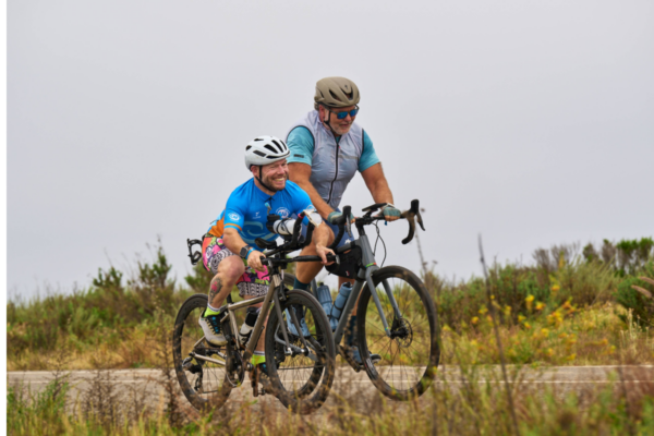An adaptive cyclist and cycling partner riding road bikes together on a paved path surrounded by coastal vegetation.