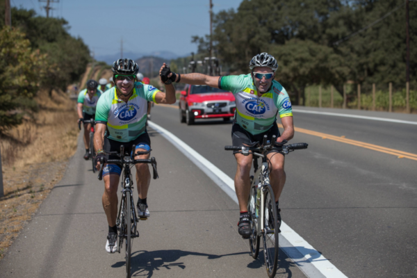 Two cyclists wearing Challenged Athletes Foundation jerseys ride on a rural road during a long-distance fundraising ride.