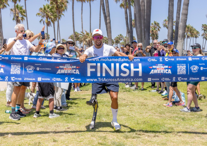 Adaptive athlete Mohamed Lahna crosses the finish line of Triathlon Across America on a running prosthesis as spectators gather at a beachside finish area in Los Angeles.