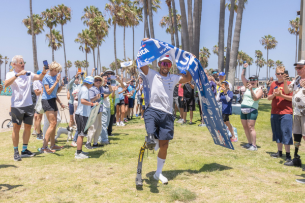 Mohamed Lahna carries a finish line banner after completing Triathlon Across America while supporters cheer at a beachside celebration in Los Angeles