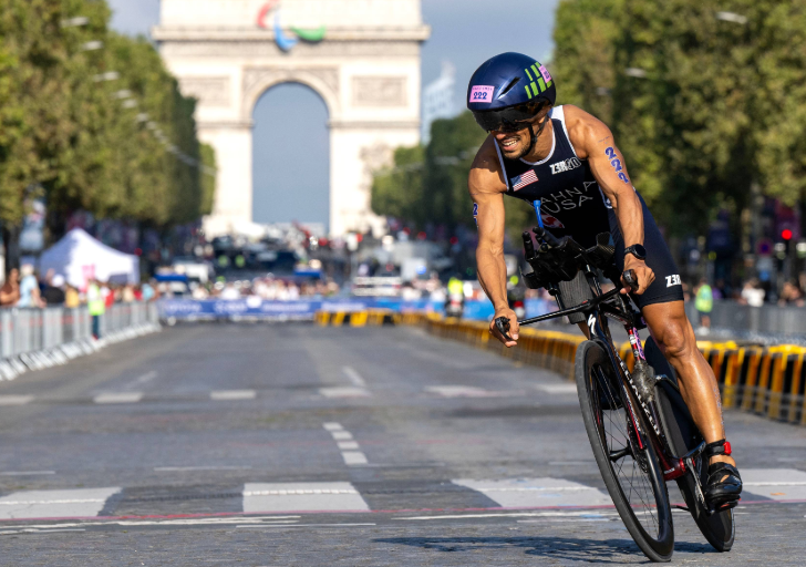 Paralympic handcyclist competing on a race course in Paris with the Arc de Triomphe visible in the background.