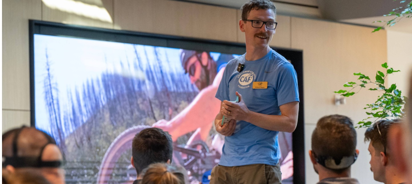 A speaker in a blue shirt presents to an engaged audience in a modern setting, with a large screen showing a cyclist in a forested landscape behind him.