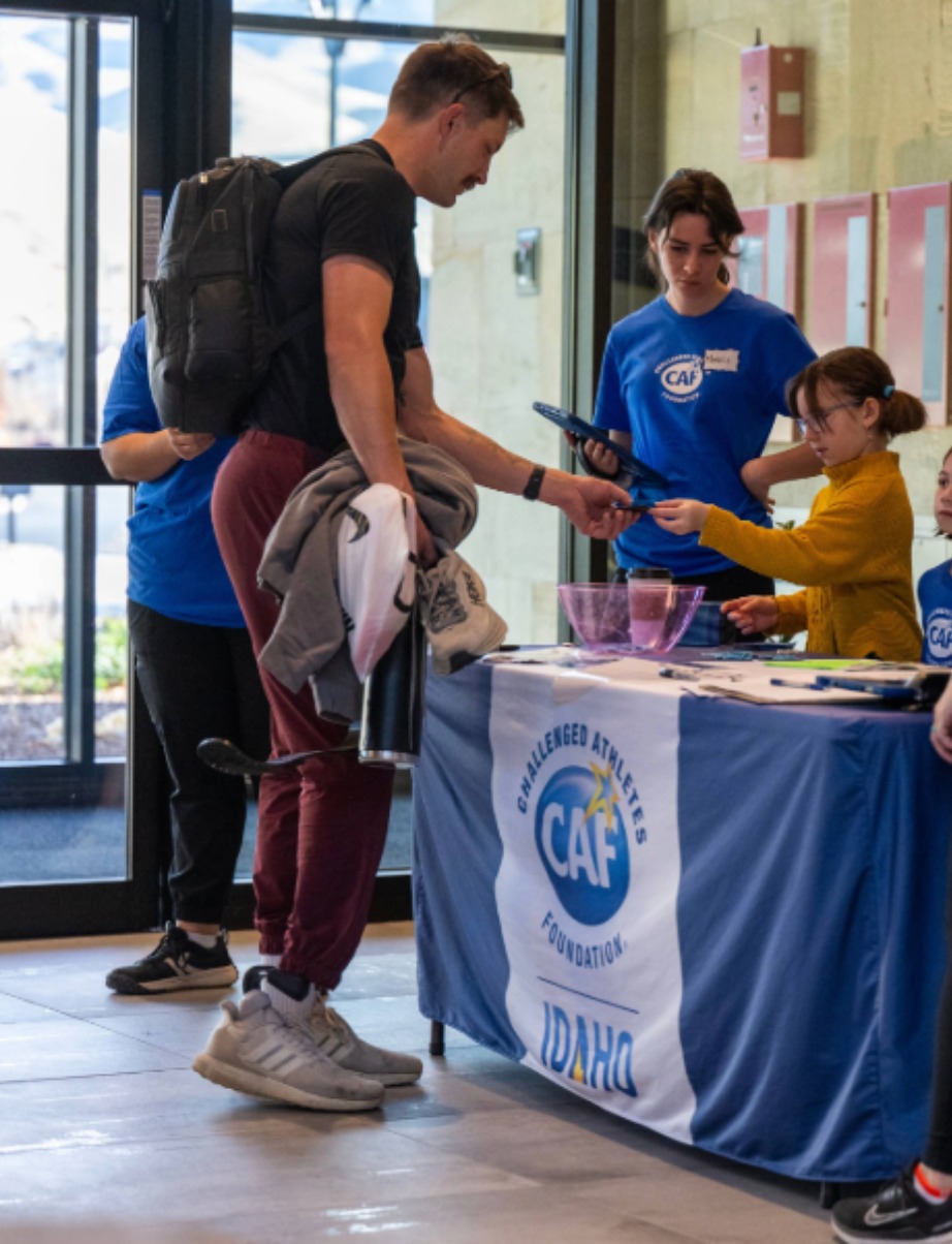 A man with a backpack interacts with individuals at a Challenged Athletes Foundation table inside a building.