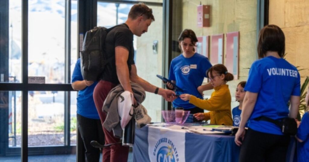 A man with a backpack interacts with individuals at a Challenged Athletes Foundation table inside a building.