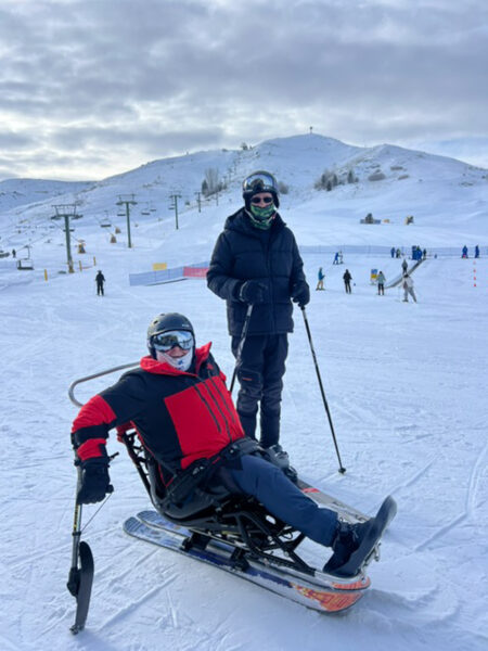 Two skiers on a snowy slope: one seated in an adaptive ski device and another standing, both in winter gear.