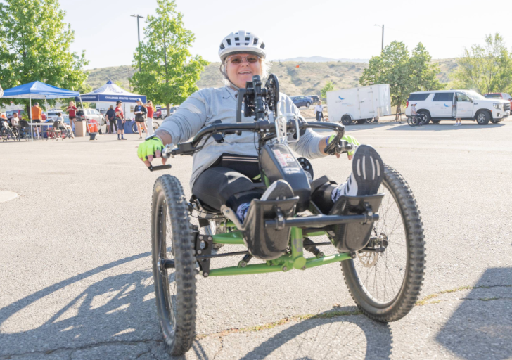 A woman on a handcycle smiles while participating in an outdoor event, surrounded by people and trees.