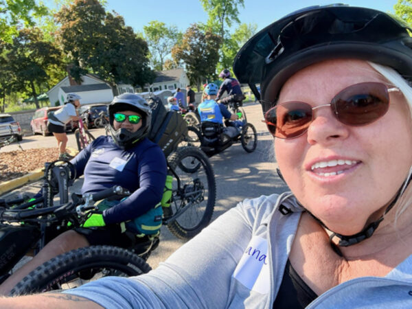 A woman takes a selfie with a man in a handcycle beside her, both smiling in an outdoor setting with other cyclists and trees in the background.
