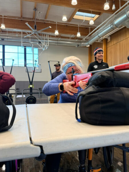 A woman aims a red rifle at a shooting range, focusing intently. Two individuals observe her, one man and one woman, in a bright, well-lit room with wooden details.