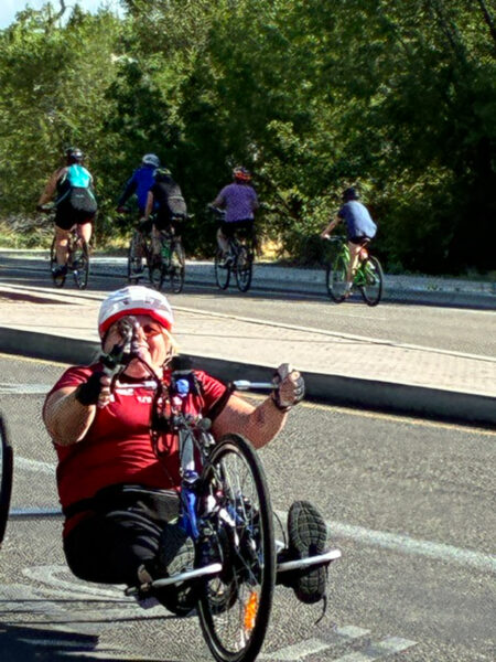 A person rides an adaptive cycle in the street, wearing a helmet and enjoying a sunny day outdoors.