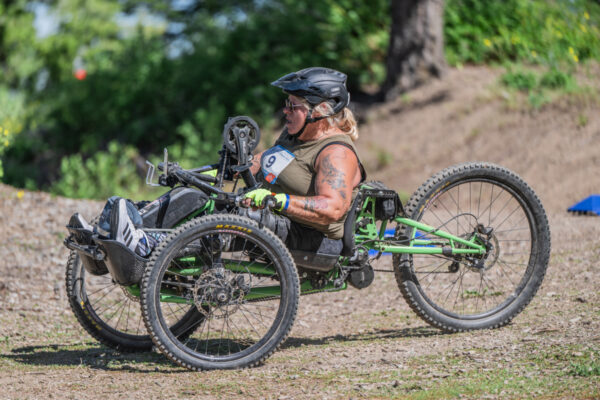 A woman with blonde hair pedals a green handcycle in an outdoor setting, wearing a black helmet and a sleeveless top. She has visible tattoos and is surrounded by greenery.