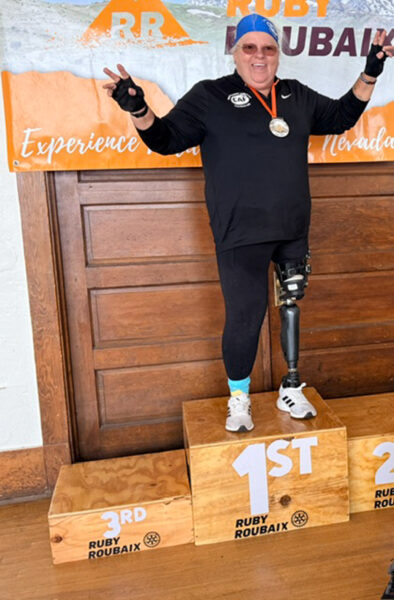 A smiling person in athletic wear stands on a first-place podium with a peace sign, displaying a silver medal. They have a prosthetic leg and are wearing a blue swim cap.