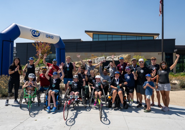 A diverse group of children and adults, some in racing wheelchairs, pose joyfully in front of a large blue arch marked "CAF IDAHO."