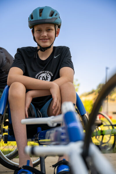 A young boy in a blue helmet sits in a wheelchair, smiling with his arms crossed. He wears a black shirt with a logo, with colorful bicycles visible in the blurred background.