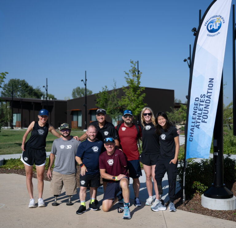 A diverse group of eight individuals posing together outdoors in athletic wear, smiling in front of a banner for the Challenged Athletes Foundation.