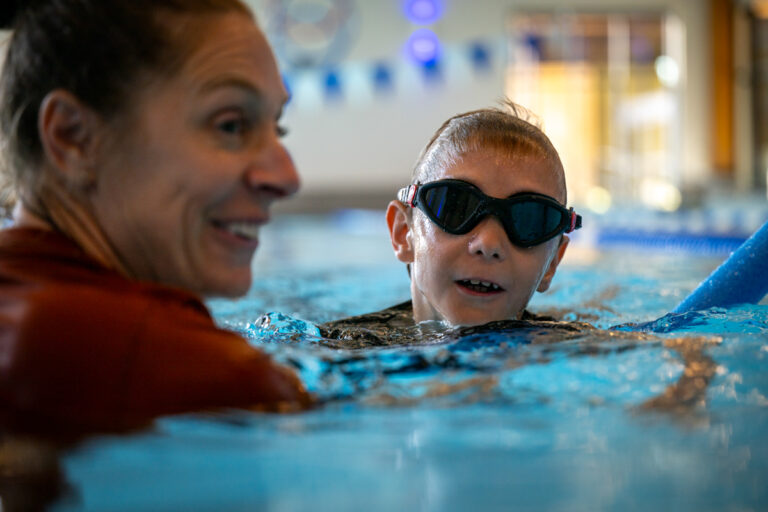 A woman and a boy in swim goggles share a joyful moment during a swimming lesson in a bright pool.