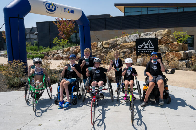 A group of six young athletes in wheelchairs, wearing gold medals, celebrate in front of an inflatable archway with CAF and Idaho logos, showcasing their triumph at a competition.