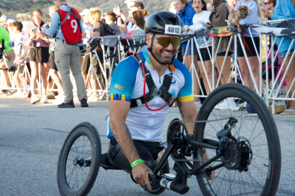 Adaptive athlete riding a handcycle during IRONMAN 70.3 Boise as spectators watch from behind course barriers.