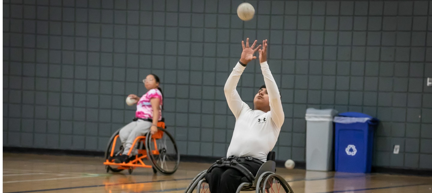 A young boy in a white shirt and dark pants reaches up from a manual wheelchair while a girl in a pink shirt watches from behind, both participating in a sports activity in a gym.