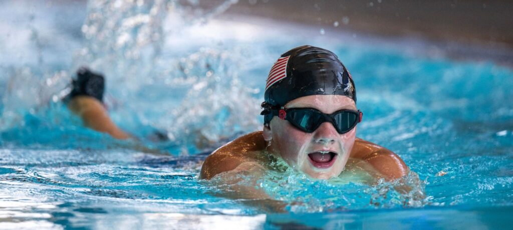 A young boy wearing a black swim cap and goggles swims in a blue pool, displaying a determined expression amidst water splashes.