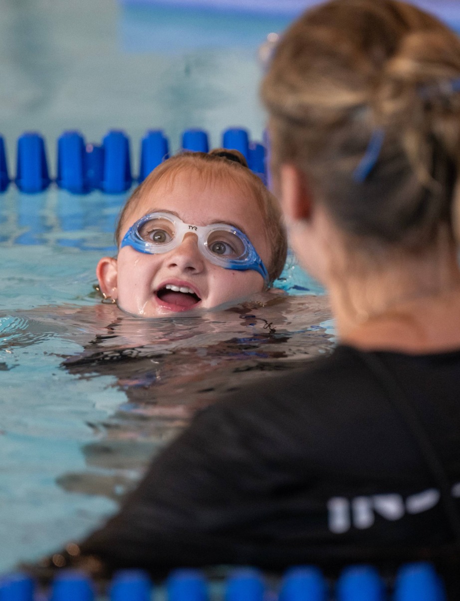 A young girl wearing goggles smiles in a swimming pool, interacting with an instructor who is partially visible to the right.