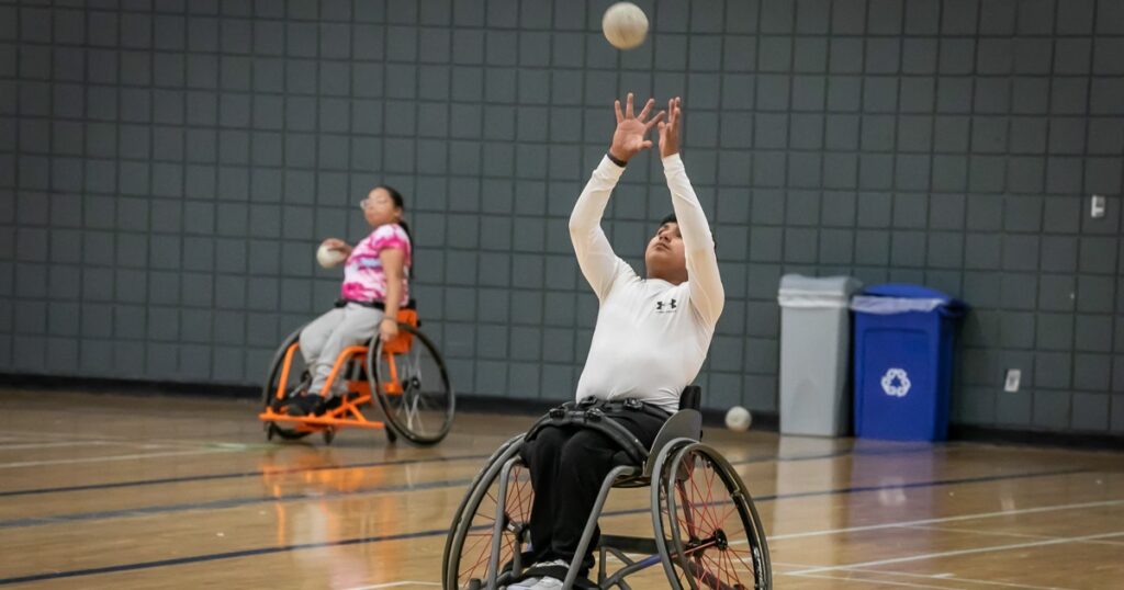 A young boy in a white shirt and dark pants reaches up from a manual wheelchair while a girl in a pink shirt watches from behind, both participating in a sports activity in a gym.