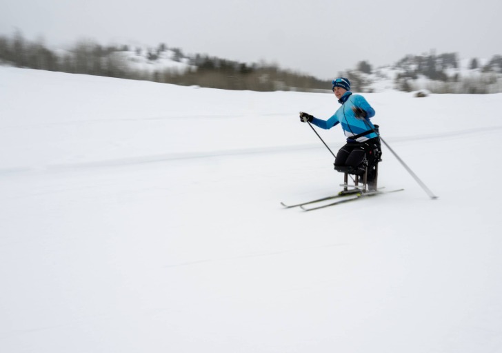 A skier in a blue jacket uses a sit-ski on a snowy landscape under a cloudy sky.