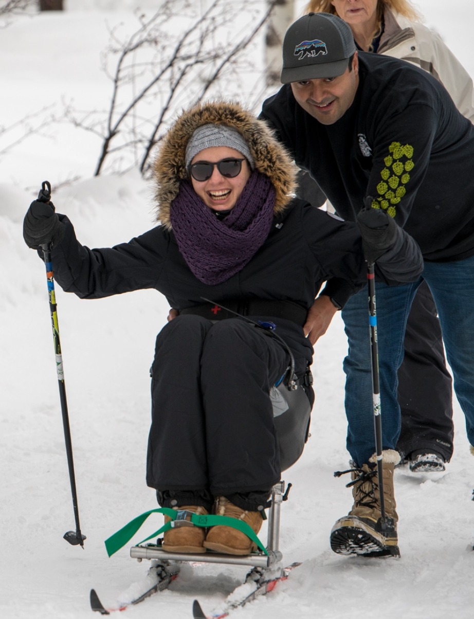A woman in a sled ski device smiles as a man pushes her through the snow; both are dressed warmly for winter.