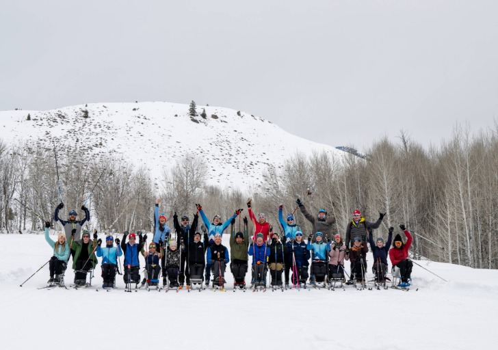 A diverse group of thirty people, some seated in adaptive ski equipment, celebrate together in a snowy landscape, with arms raised in excitement.