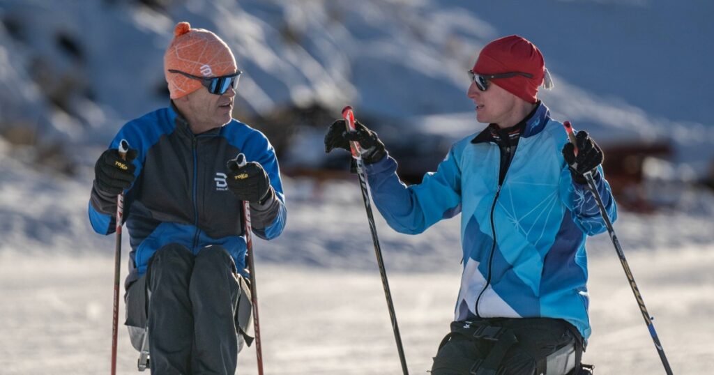 Two people in sit skis, talking to each other on the snow with a mountain backdrop