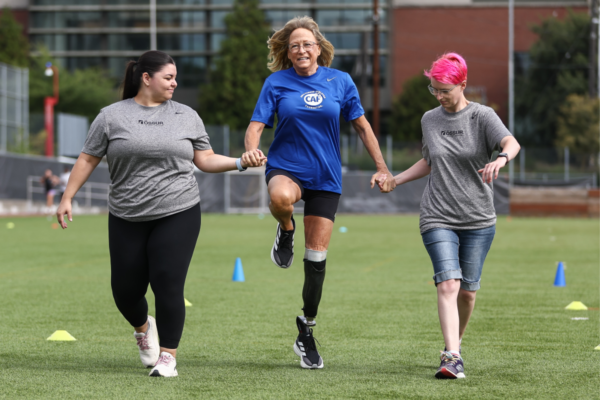 Participant using a lower limb prosthesis practices balance and movement drills with support from clinic volunteers during an adaptive running clinic.