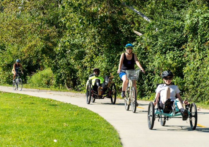 Three cyclists riding on a paved path surrounded by greenery; one on an upright bike and two on recumbent bicycles.