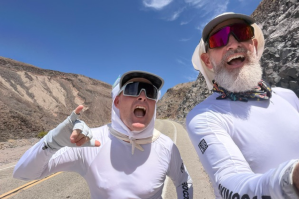 Two Badwater Ultramarathon participants wearing sun-protective gear traverse a mountain road in Death Valley during race week preparations.
