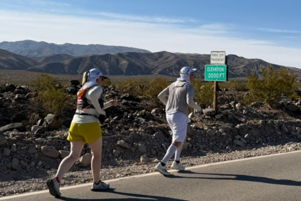 Two Badwater Ultramarathon participants travel along a remote desert roadway near an elevation marker in Death Valley's rugged landscape.