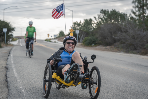 An athlete rides a yellow recumbent handcycle on a roadway with an American flag mounted behind the handcycle while another cyclist rides in the background.
