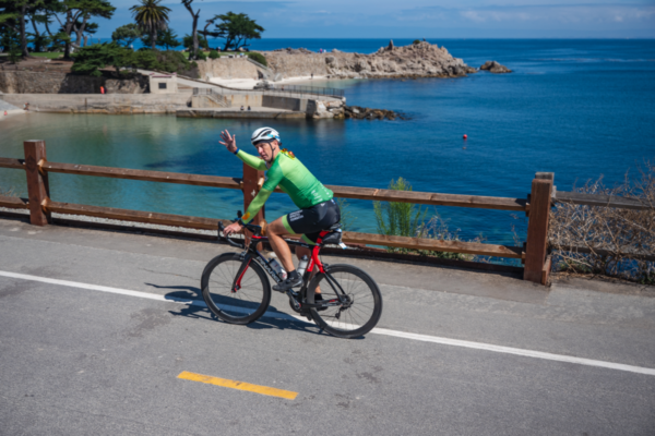 A cyclist in a green jersey rides along a coastal roadway overlooking blue water, rocky shoreline, and a small beach cove.