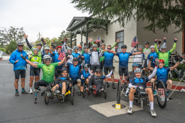 Participants gather outside a building with road bikes and recumbent handcycles during a CAF cycling event group photo.