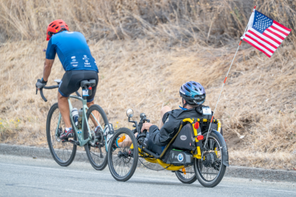 A cyclist and an athlete riding a recumbent handcycle travel along a roadway with dry grass on the roadside and an American flag attached to the handcycle.