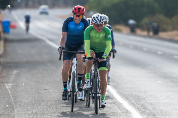 Two cyclists ride in single file on a paved road wearing blue and green CAF cycling jerseys during a group ride.