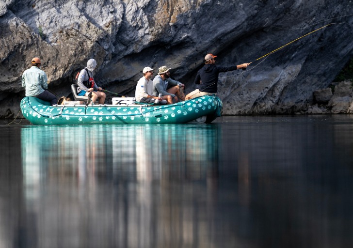 Five people in a green inflatable boat fish beside a rugged rock cliff, reflected in calm water.