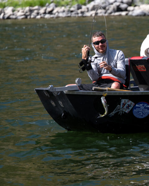 Person in a black fishing boat holds a bent fishing rod over a river while a fish hangs from the boat’s side.