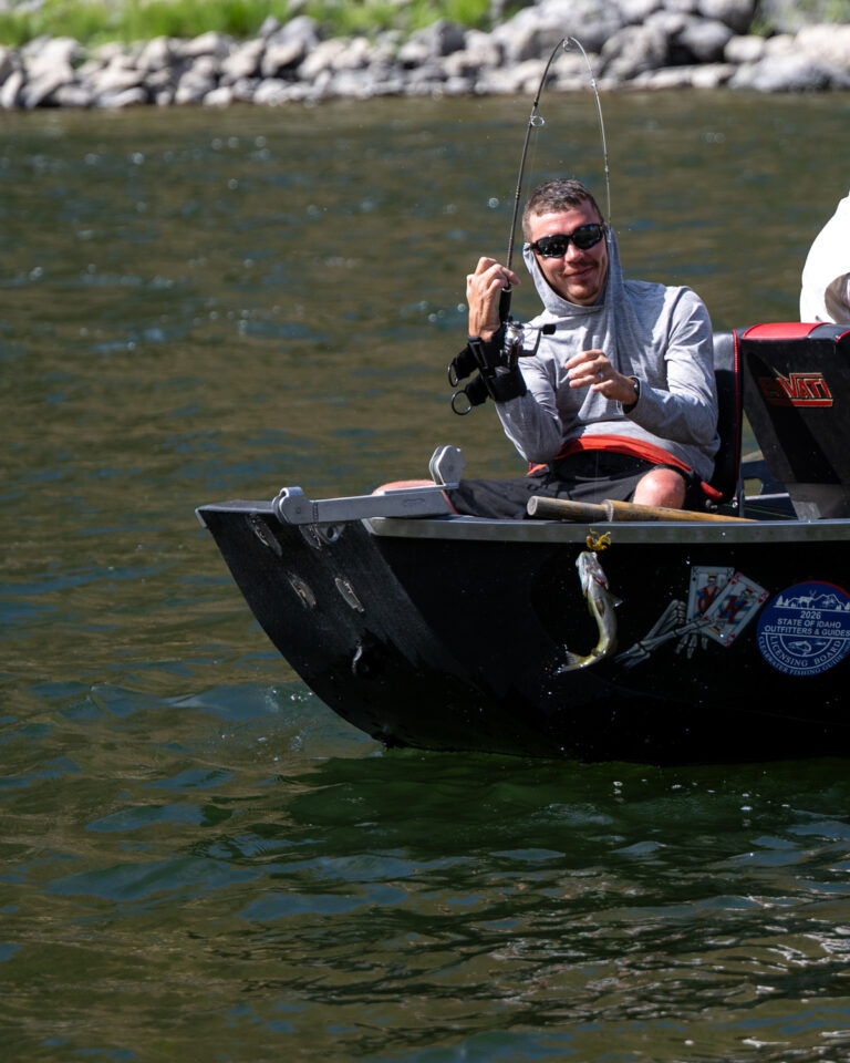 Person in a black fishing boat holds a bent fishing rod over a river while a fish hangs from the boat’s side.