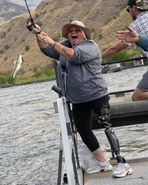 Woman with a prosthetic leg fishing from a boat as a fish hangs from her line, with dry hills in the background.